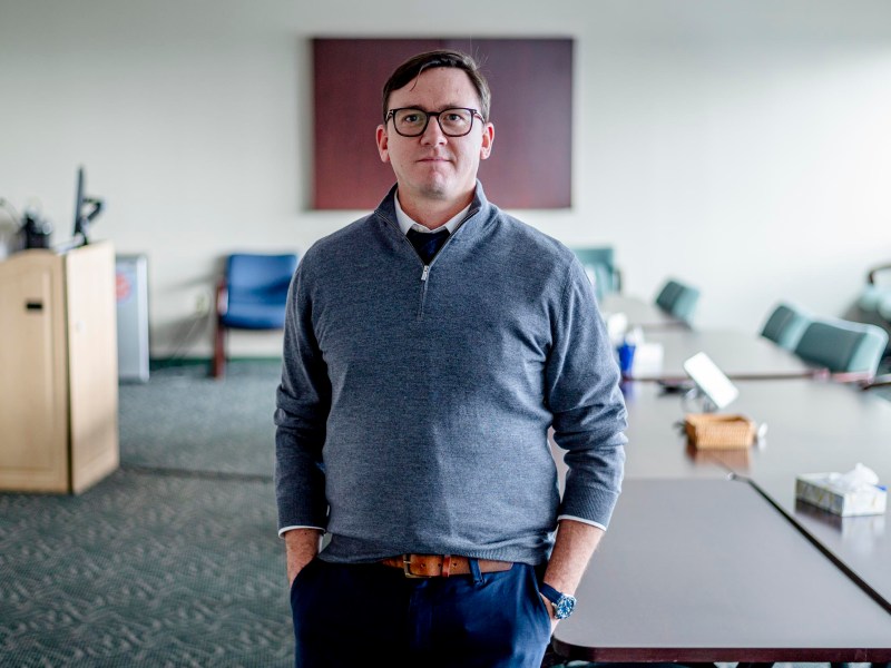 A man wearing glasses and a gray sweater stands with hands in pockets in a conference room with tables and chairs.