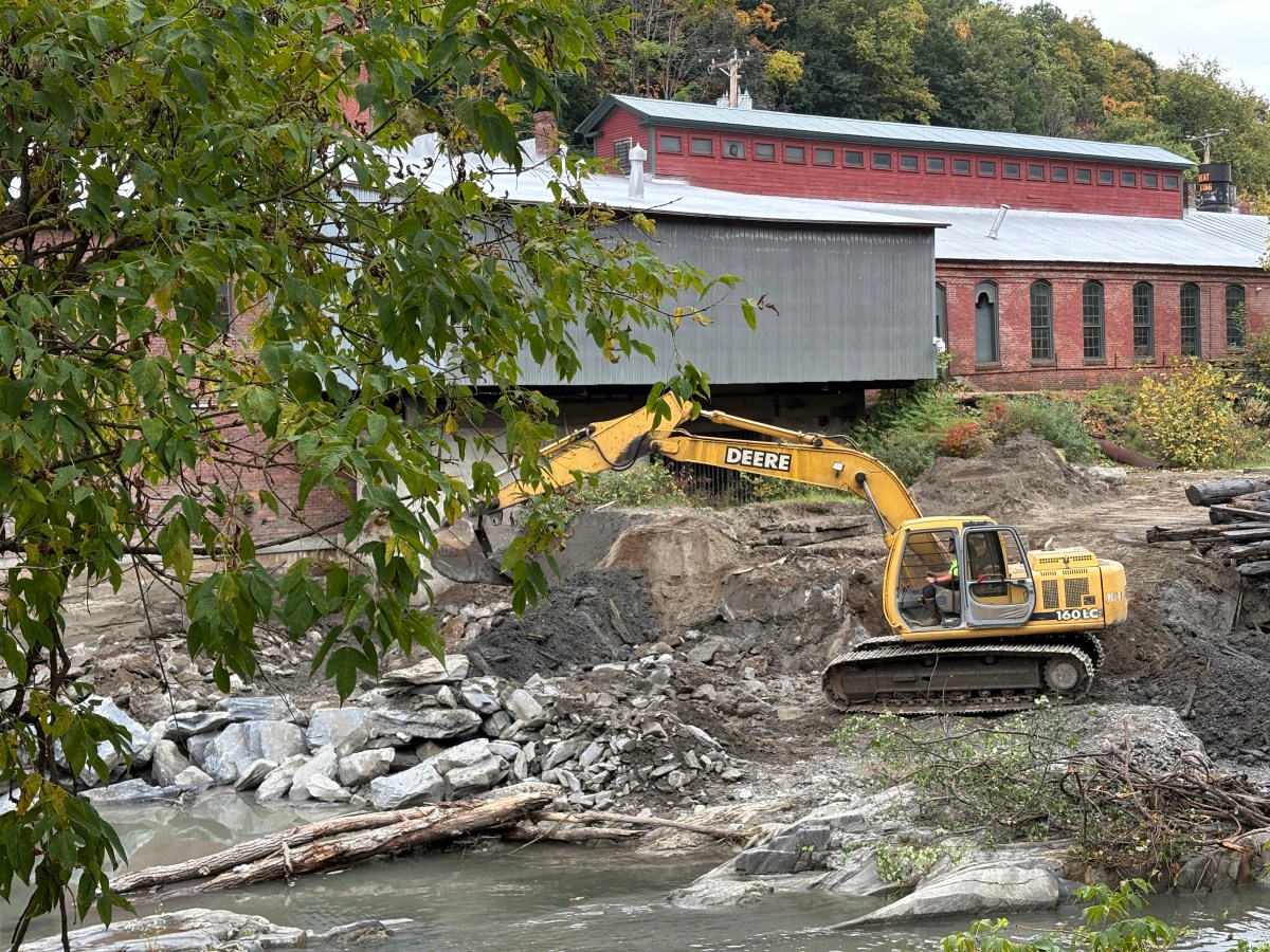 A yellow excavator moves dirt and rocks beside a river, near a covered bridge and a red brick industrial building surrounded by trees.
