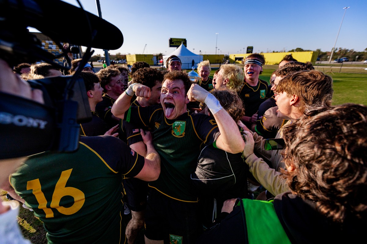A rugby player in a green and black uniform celebrates energetically with teammates on the field, surrounded by a crowd and a cameraman recording the moment.