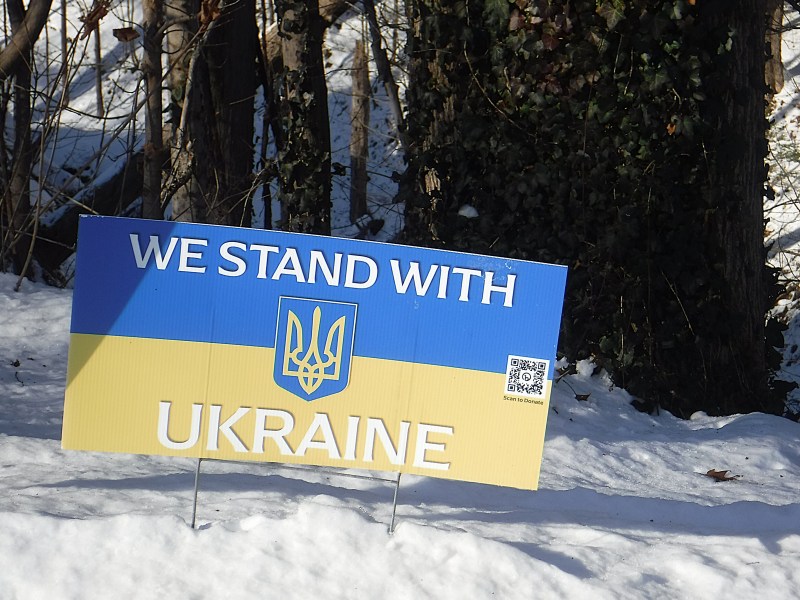 A sign in the snow reads "We Stand With Ukraine" above a yellow and blue Ukrainian flag with a trident symbol, positioned in front of trees.