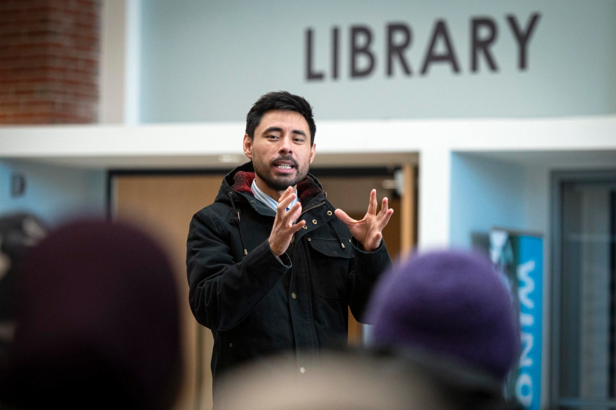 A man in a black jacket gestures while speaking to a group in front of a library entrance.