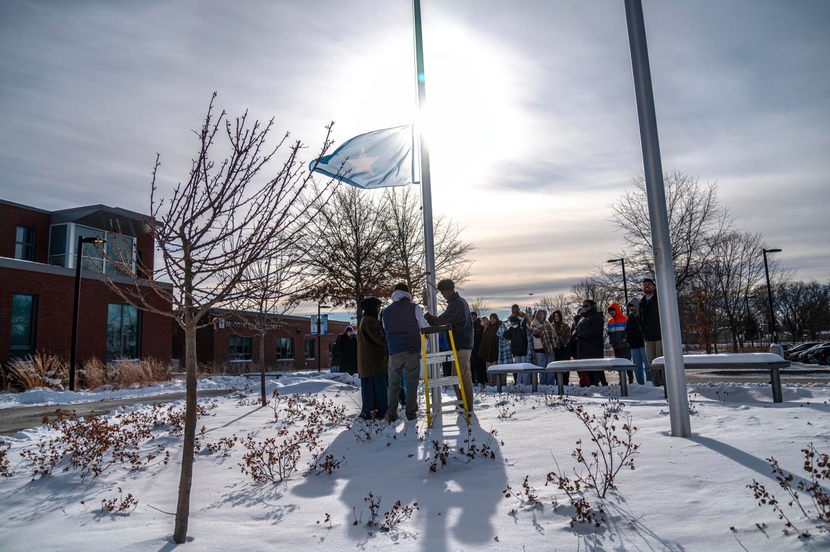 A group of people stands around a flagpole in a snowy outdoor area, raising a flag with the sun shining brightly behind them.
