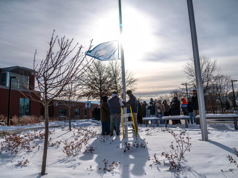 A group of people stands around a flagpole in a snowy outdoor area, raising a flag with the sun shining brightly behind them.