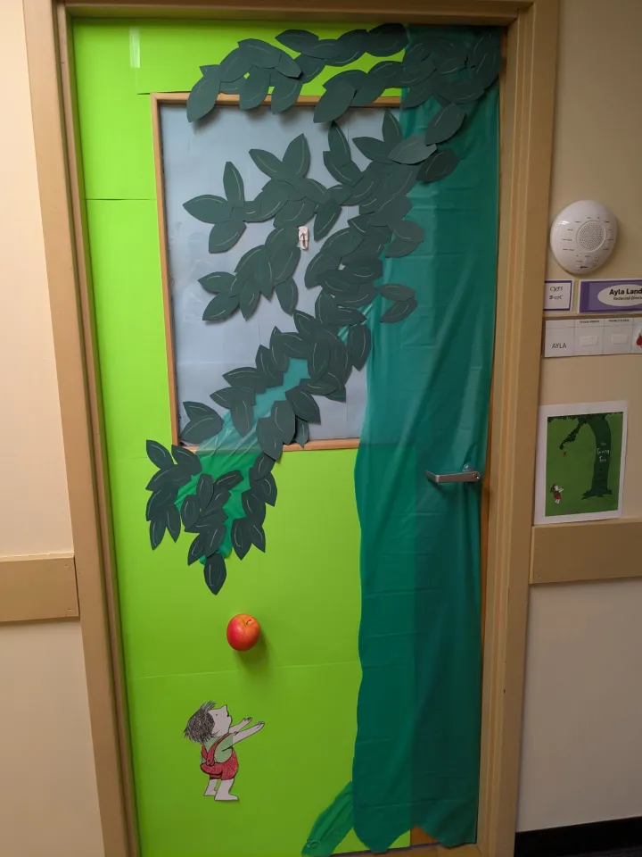 A door decorated to resemble a scene from "The Giving Tree," featuring a tree with green leaves, an apple, and a child standing below, with paper cutouts and colored paper.