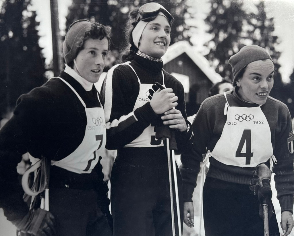 Three female cross-country skiers wearing numbered bibs and winter gear stand together outdoors at the 1952 Oslo Winter Olympics.