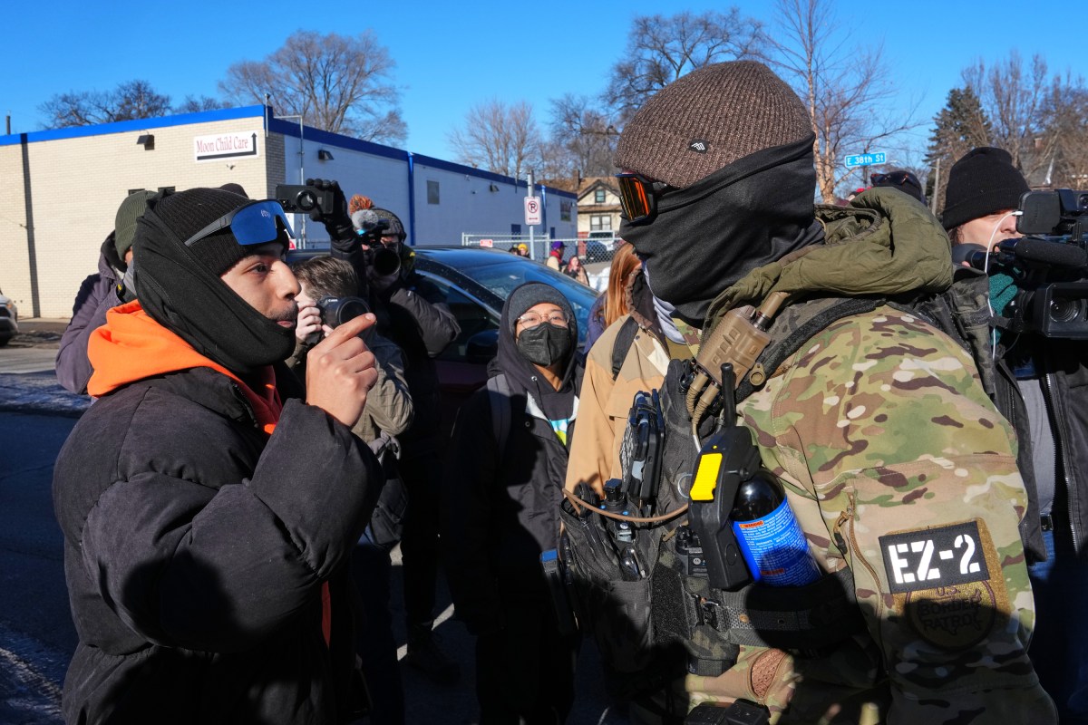 A man gestures and speaks to a camouflage-clad law enforcement officer during a winter street protest, while others observe and media capture the scene.