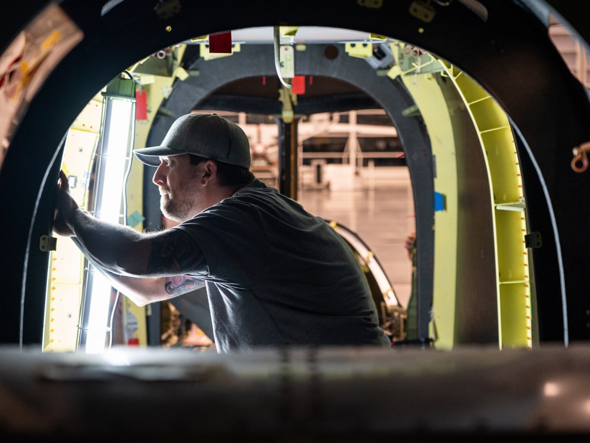 A man in a cap works inside the circular frame of an aircraft fuselage, inspecting or assembling components in a well-lit industrial setting.