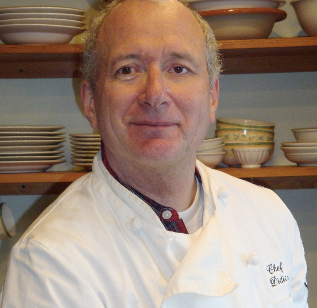 A chef in a white coat stands in front of shelves filled with stacked dishes and bowls, looking at the camera and smiling slightly.