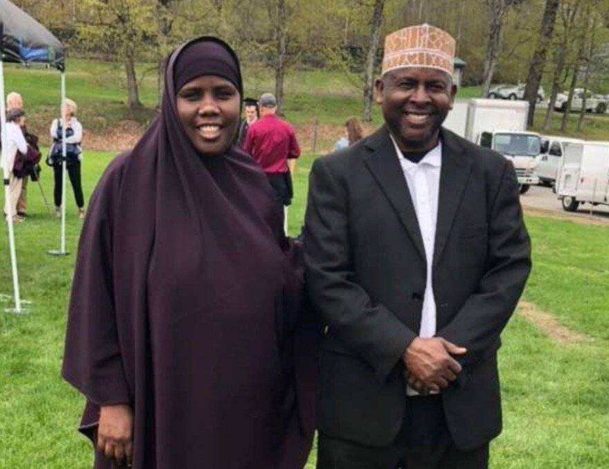 Two adults stand side by side outdoors on grass, smiling at the camera. The woman wears a dark hijab and the man wears a suit and patterned cap. Other people and trees are in the background.