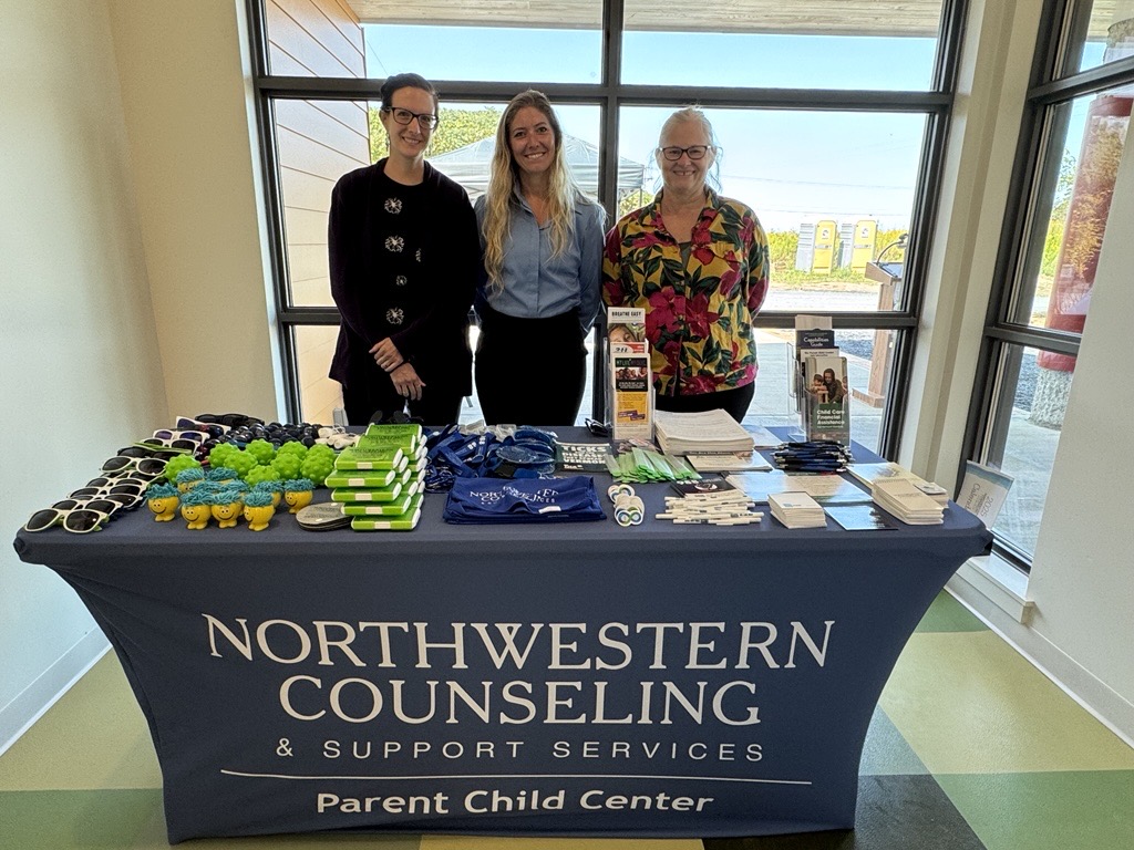 Three people stand behind a table covered with informational materials and promotional items for Northwestern Counseling & Support Services Parent Child Center.