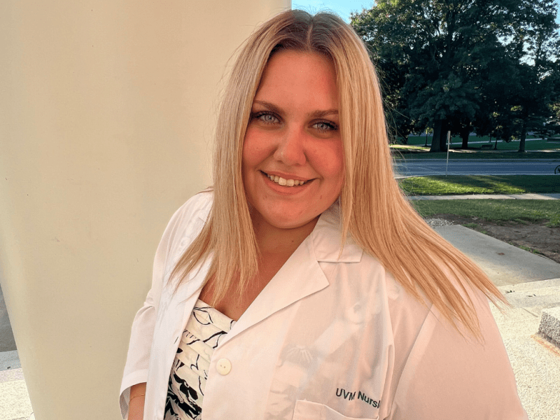 A woman with long blonde hair, wearing a white lab coat, stands outside near a column and smiles at the camera. Trees and a road are visible in the background.