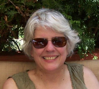 A smiling older woman with short gray hair and sunglasses sits in front of green foliage, wearing a sleeveless top.