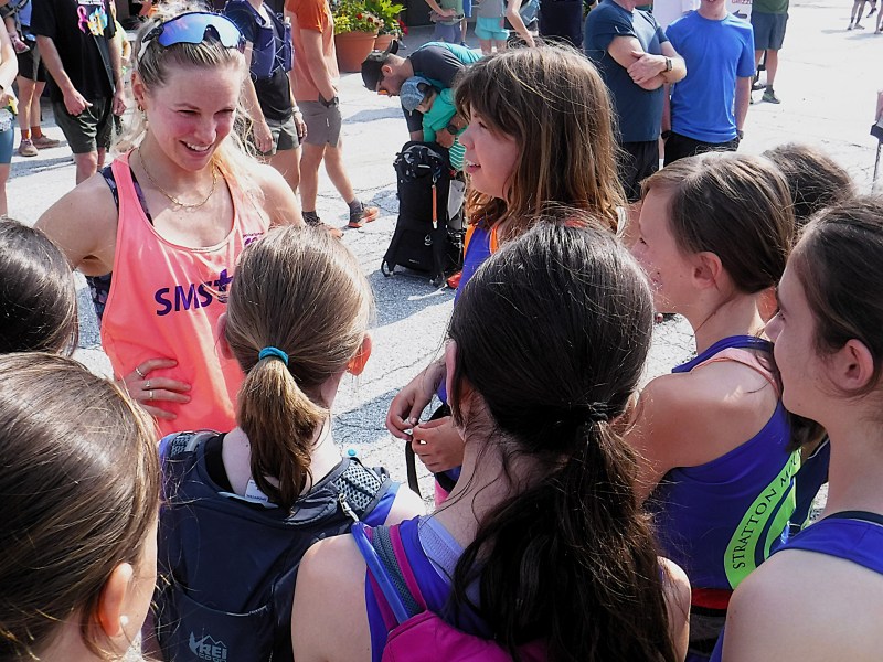 A woman in athletic wear stands smiling and talking with a group of young girls outdoors at what appears to be a sporting event. Other people are visible in the background.