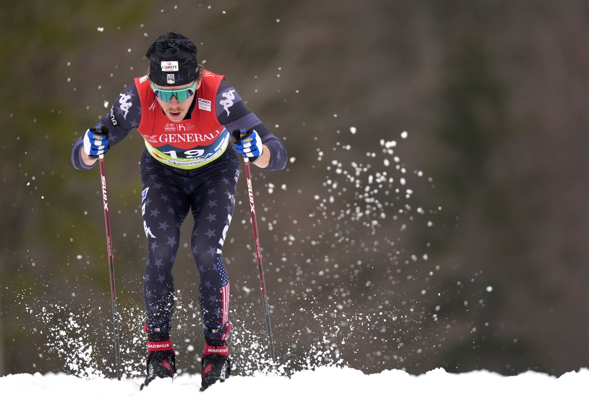 A cross-country skier wearing a race bib and USA-themed gear uses ski poles while moving through snow, with snow spray visible around their skis.
