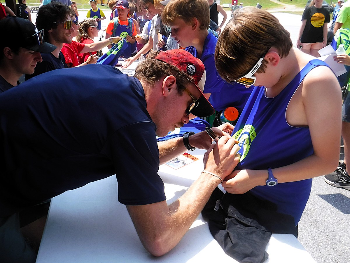 An adult signs a young person's shirt at an outdoor event while others wait in line.