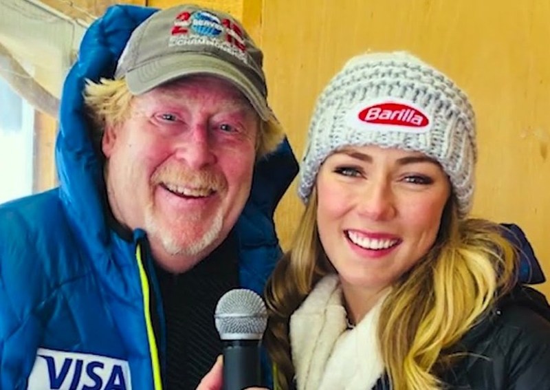 Two people in winter clothing, one holding a microphone. Both are smiling and wearing jackets and branded hats. They appear to be indoors.
