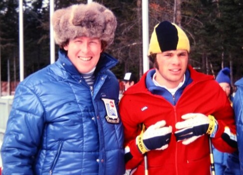 Two men outdoors in winter gear; one wears a blue puffy jacket and fur hat, the other a red jacket, yellow-black hat, and ski gloves, holding ski poles. Trees in the background.