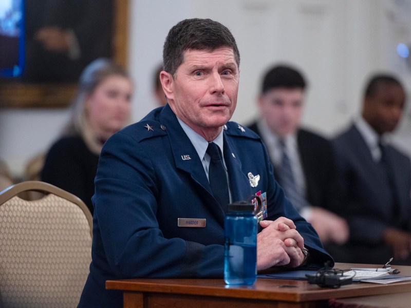 A uniformed military officer sits at a table with a blue water bottle, while several people sit in the background during a formal meeting.