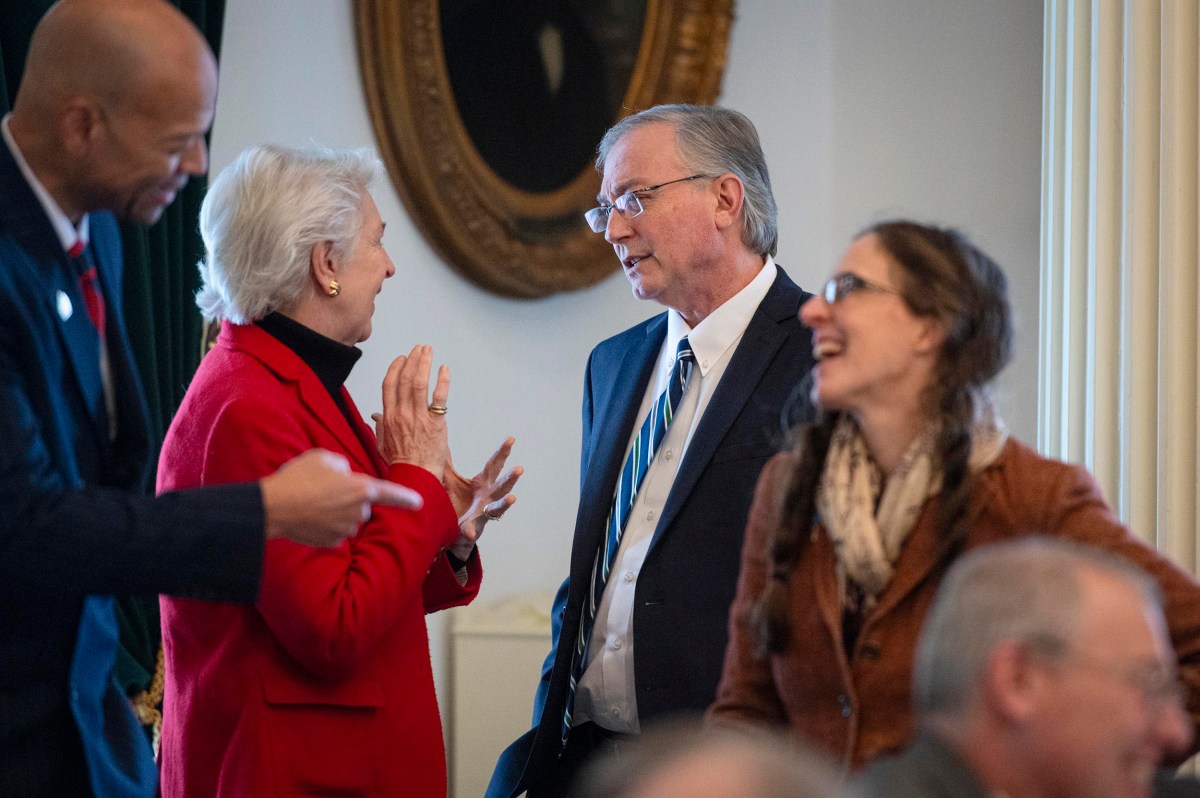 Four adults converse in a formal indoor setting; two are engaged in discussion while others stand nearby, one smiling and another holding a pen.