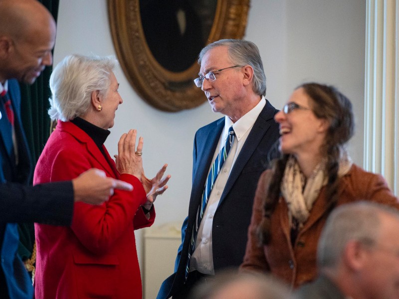 Four adults converse in a formal indoor setting; two are engaged in discussion while others stand nearby, one smiling and another holding a pen.