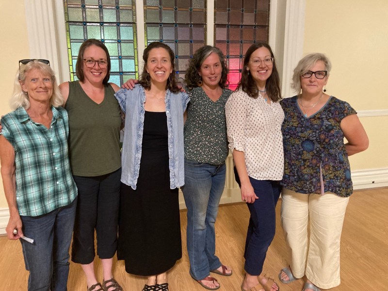 Six women stand side by side indoors in front of a window with colored glass panes, posing and smiling for a group photo.