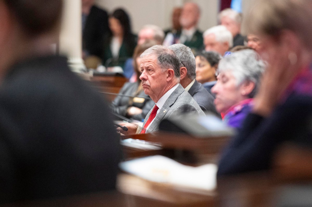A man in a gray suit and red tie sits among other people in a crowded meeting or legislative chamber, appearing focused.