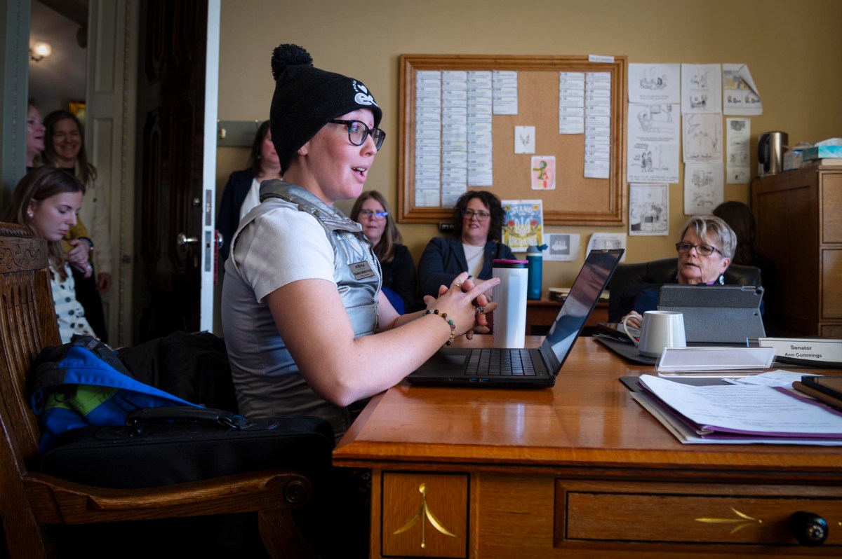A person wearing a knit hat speaks at a desk with a laptop in an office setting, while several others listen attentively.