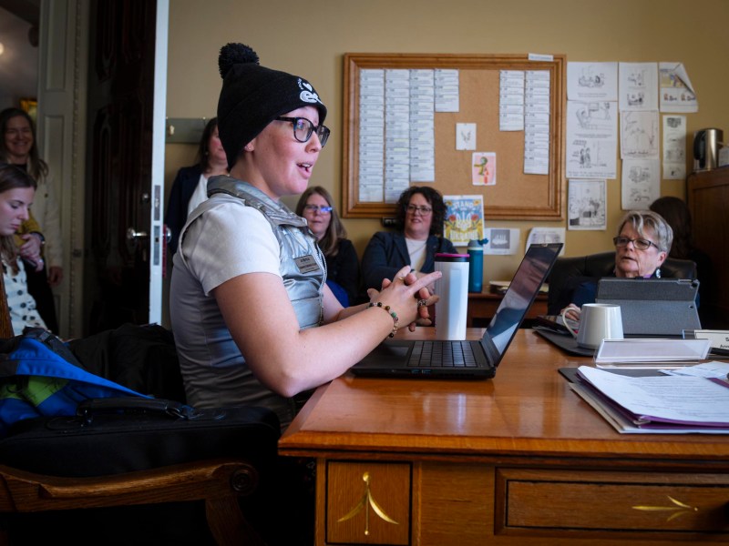 A person wearing a knit hat speaks at a desk with a laptop in an office setting, while several others listen attentively.