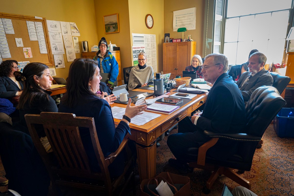 Eight people sit around a crowded office desk engaged in discussion; documents, mugs, and folders are visible, with sunlight streaming through a large window.