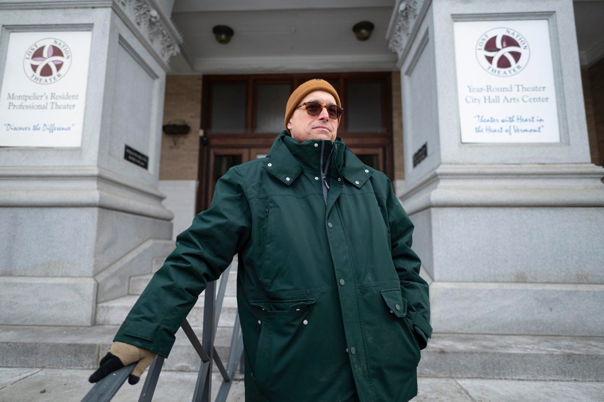 A person in a green coat and brown hat stands in front of a theater building with signs on either side of the entrance columns.