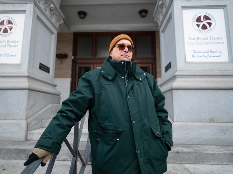 A person in a green coat and brown hat stands in front of a theater building with signs on either side of the entrance columns.