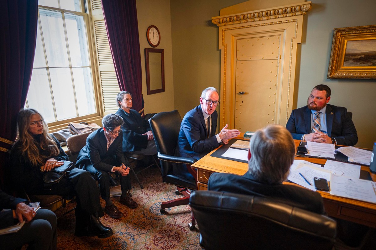 A group of people in business attire have a meeting around a desk in an office with documents, a window, and framed artwork on the wall.