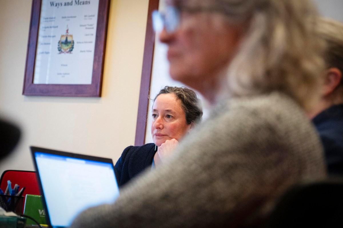 A woman sits attentively at a table during a meeting, with a blurred person and an open laptop in the foreground. A framed "Ways and Means" sign hangs on the wall behind her.