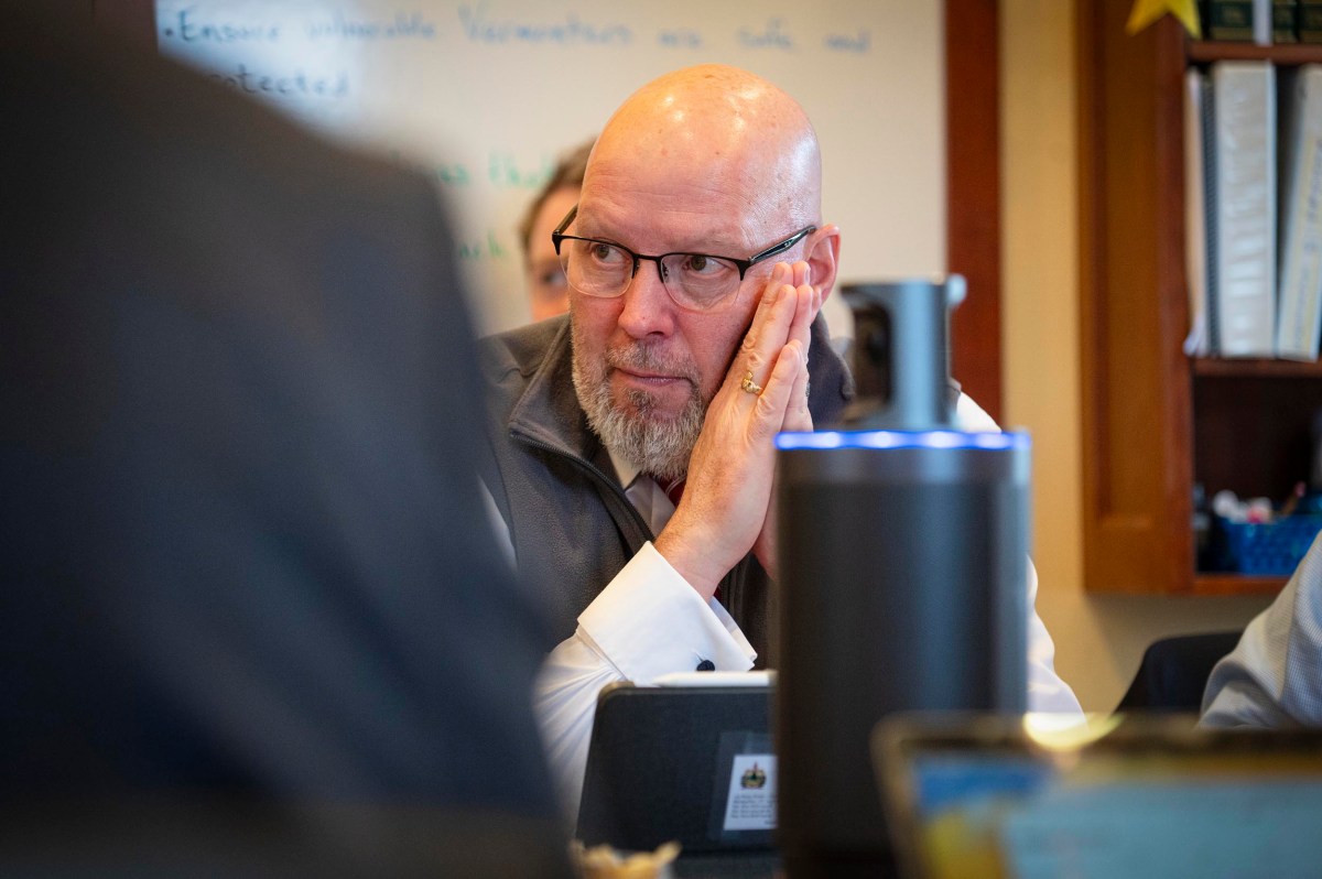 Bald man with glasses and a white shirt rests his head on his hand while seated at a table in an office or meeting room.