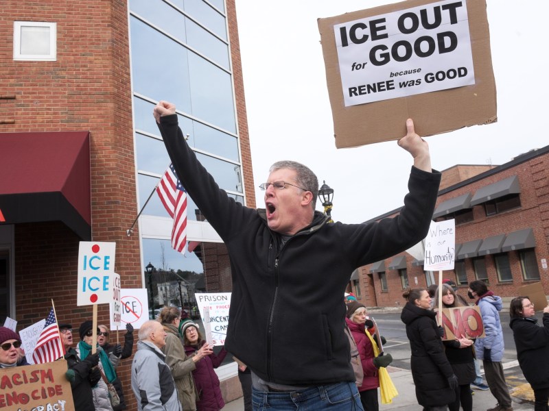 A man holds a sign reading "ICE OUT FOR GOOD because RENEE was GOOD" and shouts at a protest, with other people holding signs in the background.