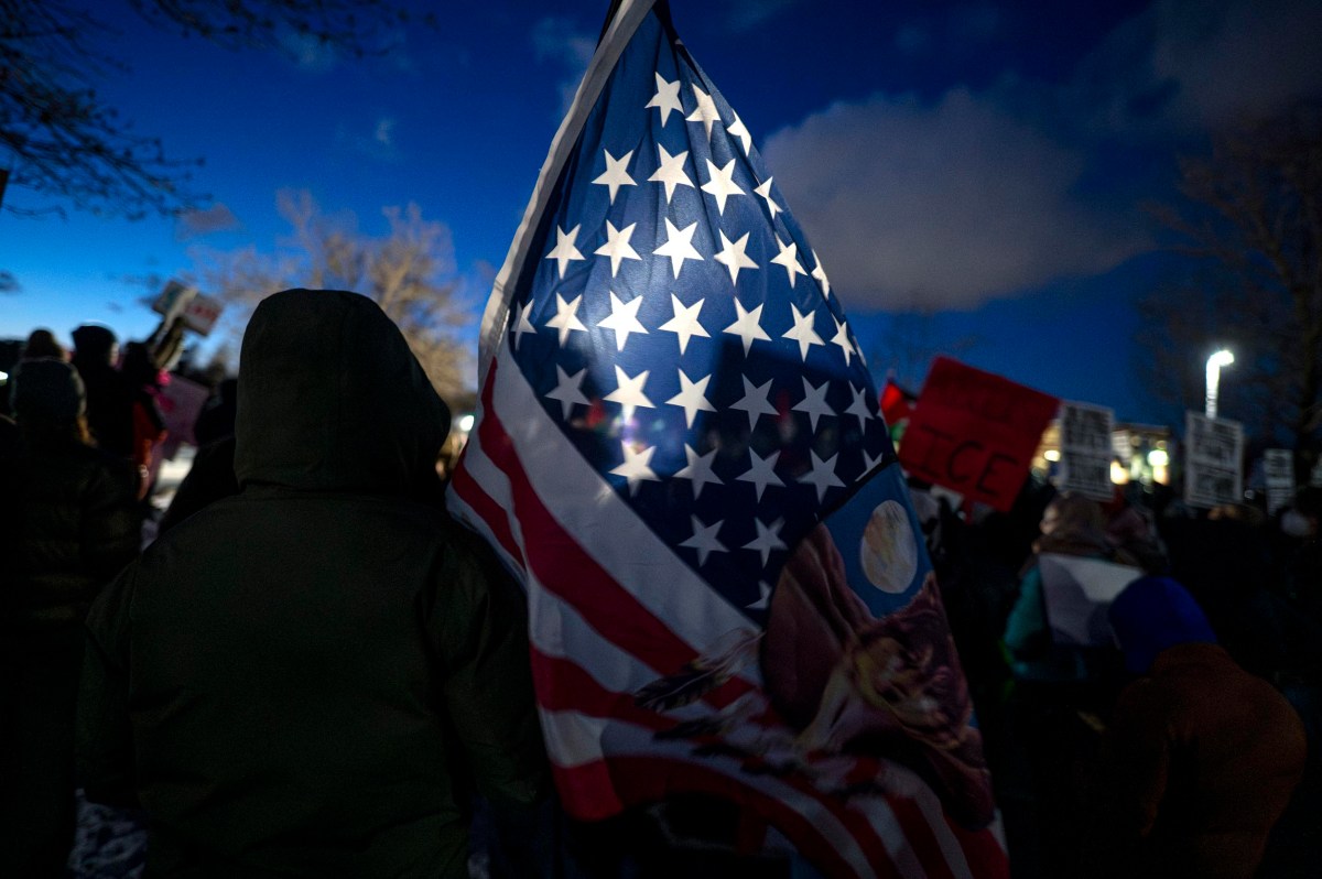 A group of people stand outdoors at dusk, with one person holding a large American flag illuminated by light. Protest signs are visible in the background.