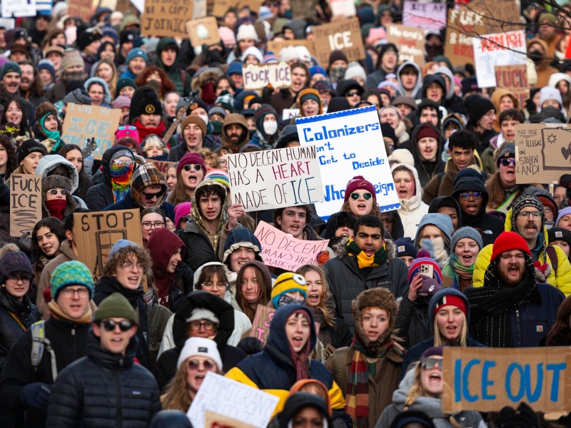 A large group of people at a protest hold signs and banners with messages related to climate change and anti-colonialism. Many are dressed in winter clothing.