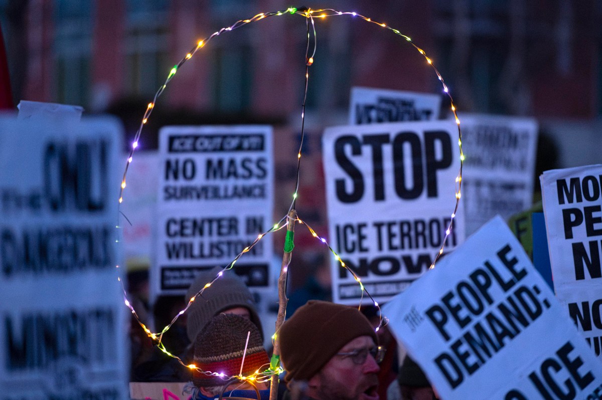 Protesters hold signs and a large illuminated peace symbol at a demonstration against ICE and mass surveillance.