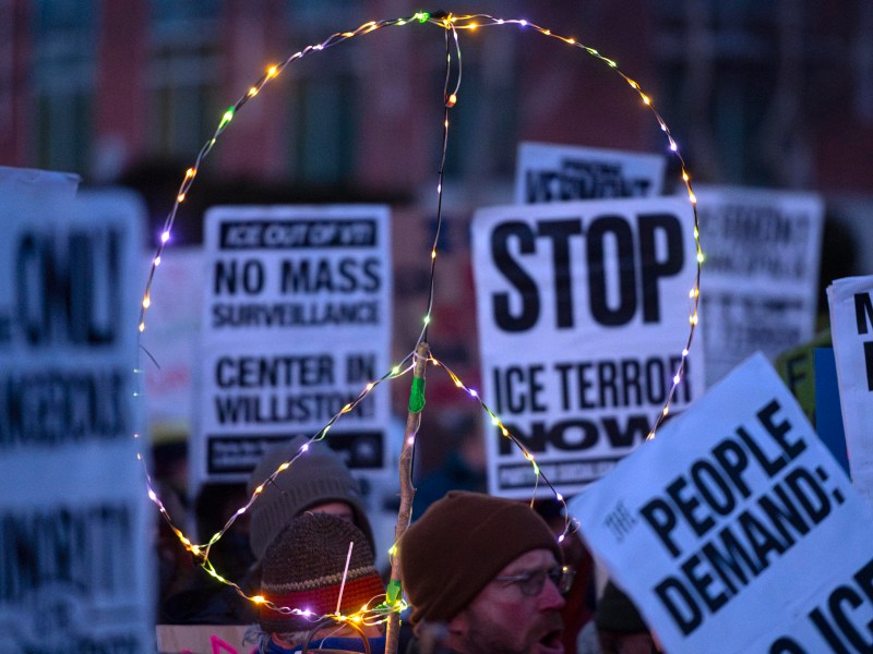 Protesters hold signs and a large illuminated peace symbol at a demonstration against ICE and mass surveillance.