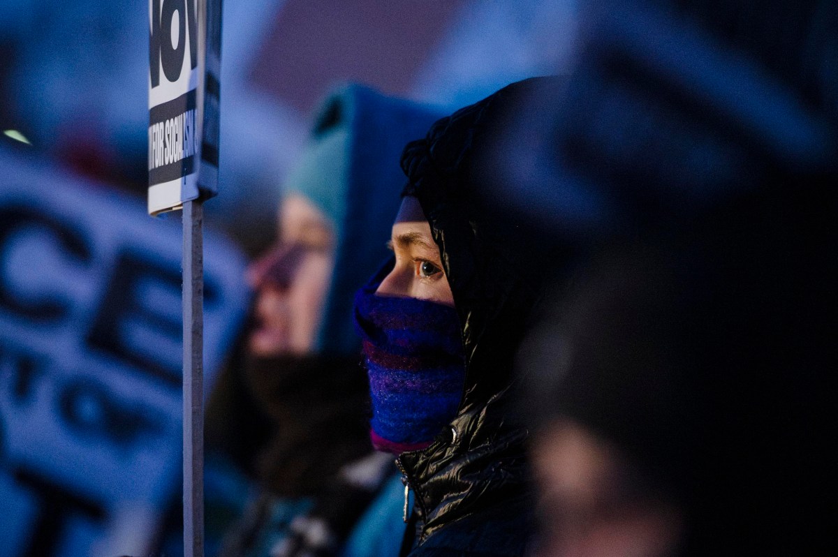 Person wearing a hooded jacket and a face covering stands among a group of people holding protest signs in a dimly lit outdoor setting.