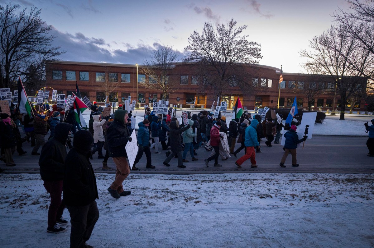 A group of people march on a snowy street, holding signs and flags during a protest near a brick building at dusk.
