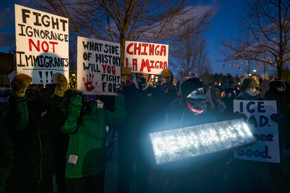 A group of protesters hold signs at night with messages such as "FIGHT IGNORANCE NOT IMMIGRANTS," "CHINGA LA MIGRA," and a lit sign reading "DEMOCRACY.