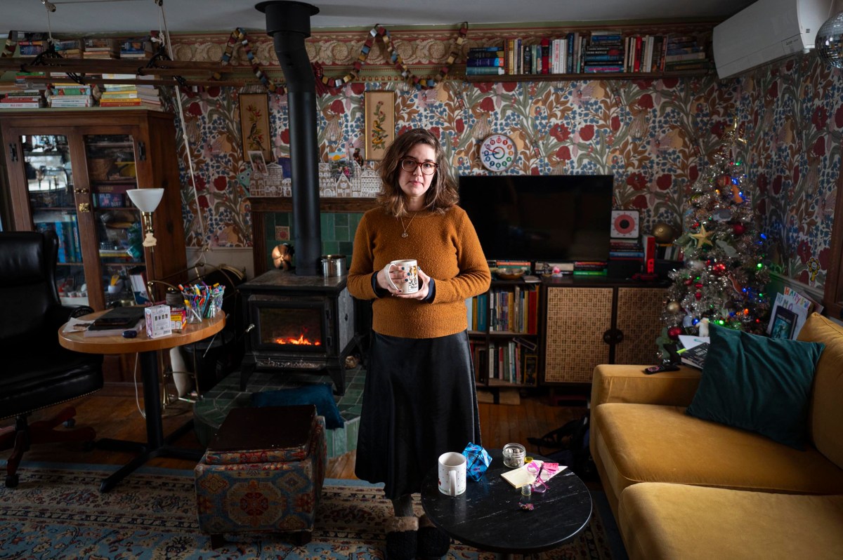 A woman stands holding a mug in a cozy, cluttered living room with patterned wallpaper, a wood stove, bookshelves, a TV, and a small decorated Christmas tree.