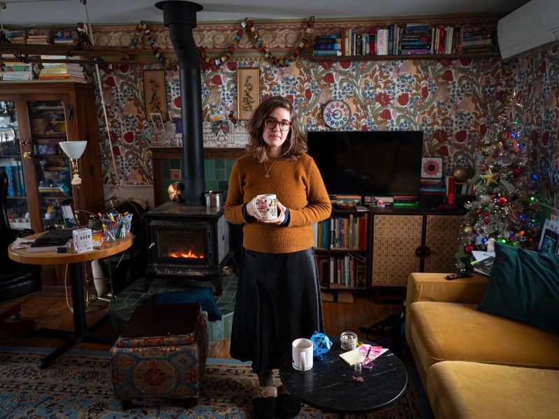 A woman stands holding a mug in a cozy, cluttered living room with patterned wallpaper, a wood stove, bookshelves, a TV, and a small decorated Christmas tree.