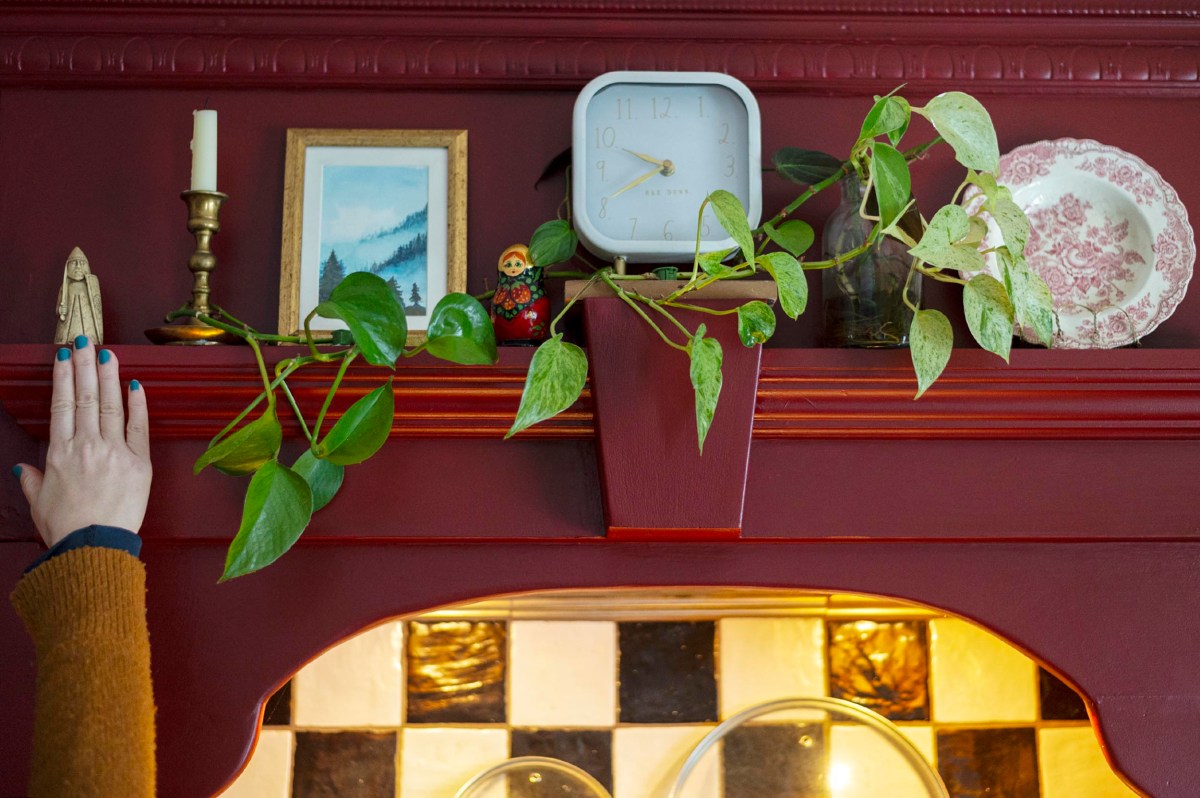 A red mantle with a plant, clock, framed picture, candle, small doll, and plate; a hand touches a wooden figurine on the left. Black and white tiles are visible below.