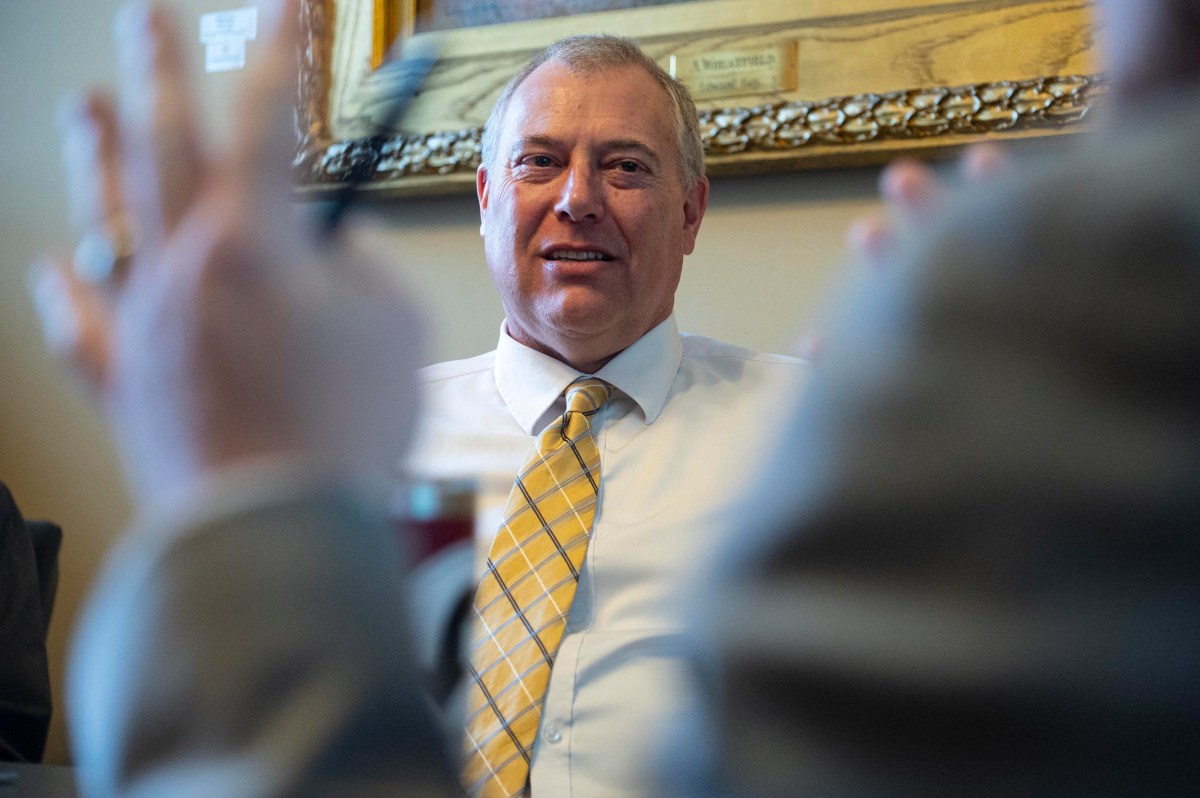 A man in a white shirt and yellow patterned tie sits at a table, with blurred hands visible in the foreground and a framed picture on the wall behind him.