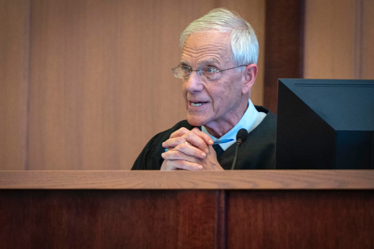 An older male judge in a black robe sits behind a bench, clasping his hands and speaking in a courtroom.