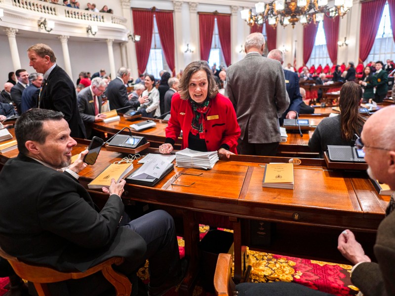 People converse and prepare materials in a large, formal legislative chamber with red curtains and wooden desks.