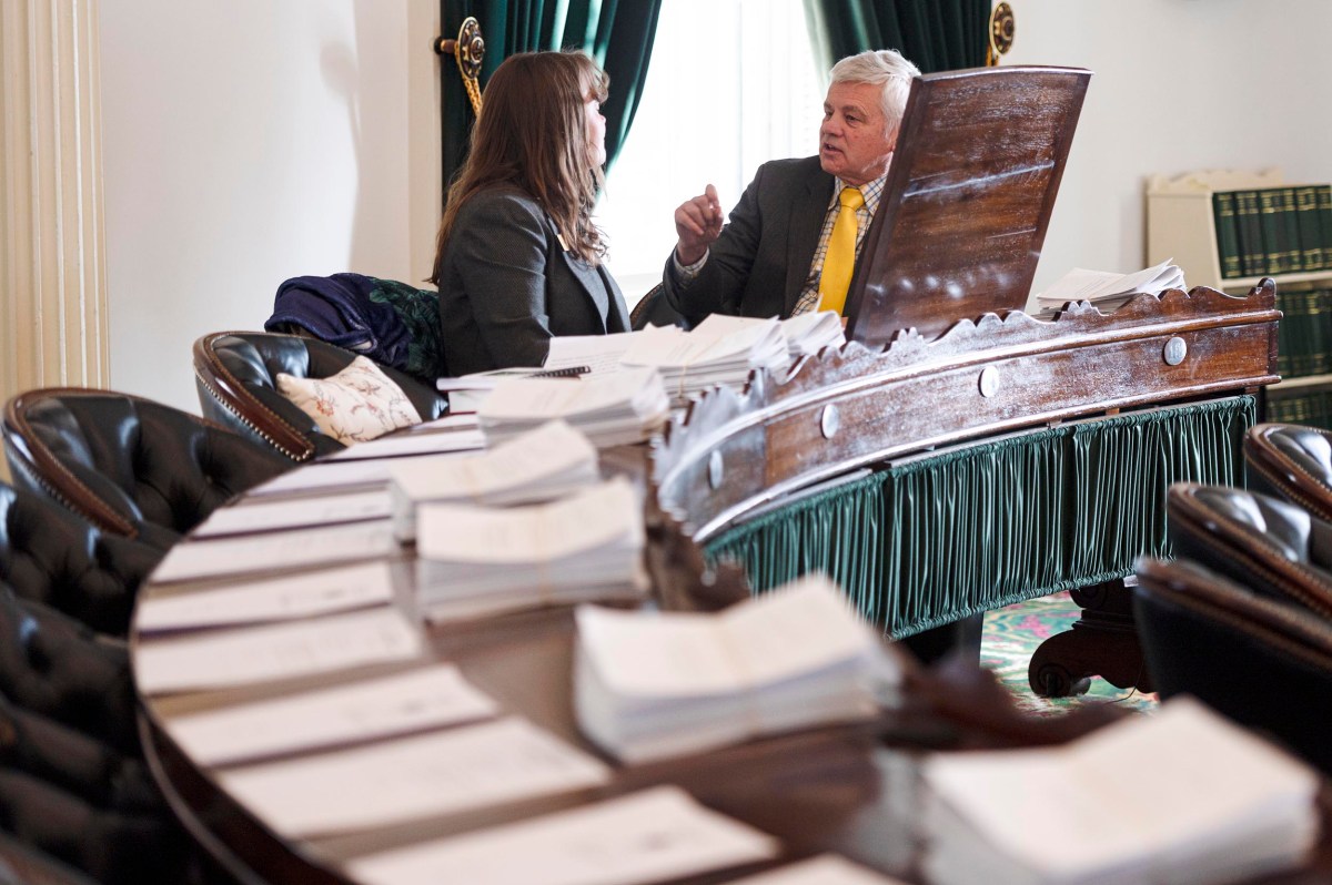 Two people in business attire have a discussion at a wooden conference table covered with stacks of documents in a formal room.