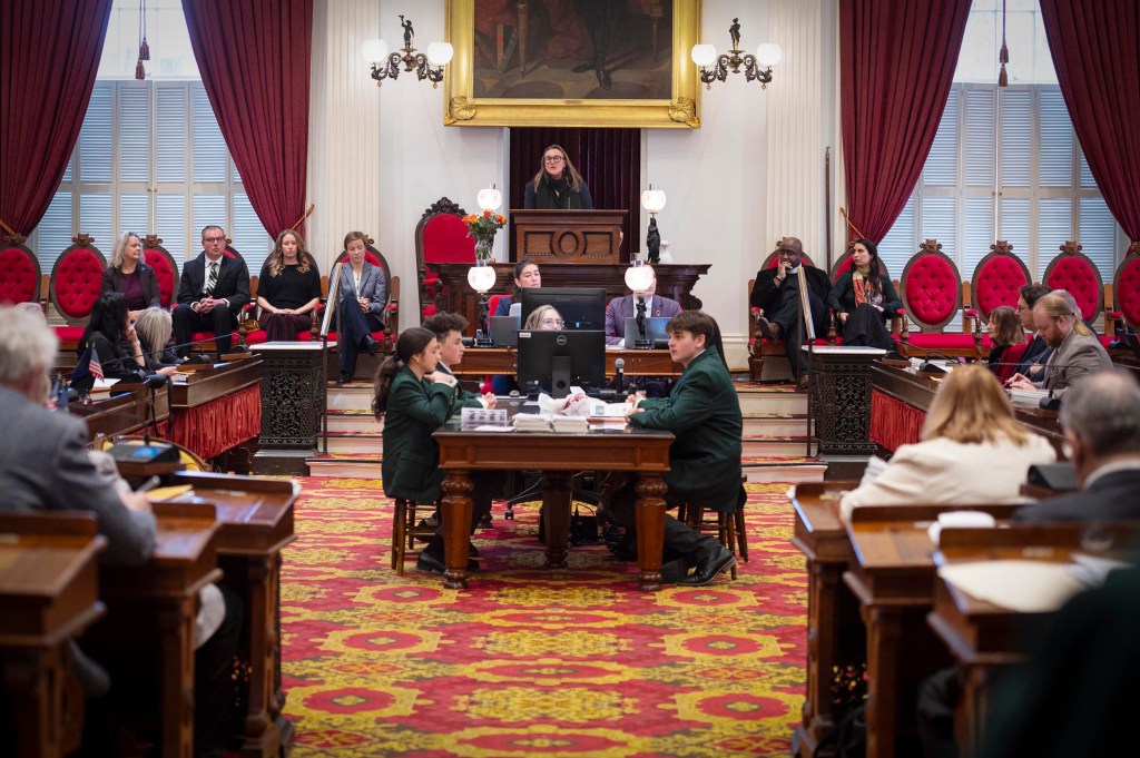 A legislative chamber with officials seated at desks, a speaker at the podium, and others seated behind; red carpets and drapes decorate the room.